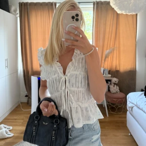 Vit blus med volang och knyt - Supersöt vit blus med volangdetaljer och knyt framtill. Blusen är ärmlös med lätt rynkade axlar och har en luftig, romantisk känsla. Perfekt till jeans eller kjol för en fräsch look. Tunt bomullstyg med diskret mönster.