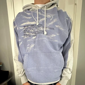 Helmut Lang hoodie i blått och grått - Säljer en hoodie från Helmut Lang med coolt blått och grått färgblocksmönster och tryck på både fram- och baksida. Tröjan har huva med snörning, känguruficka och är tillverkad i mjuk bomull. Perfekt för dig som gillar streetwear och vill sticka ut.