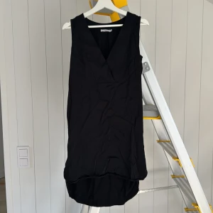 Svart ärmlös klänning från Wera - Stilren svart ärmlös klänning från Wera med v-ringning och lätt rundad nederkant. Modellen är rak och har en enkel, modern look som passar perfekt till sneakers eller boots. Tunt, mjukt tyg som faller snyggt.