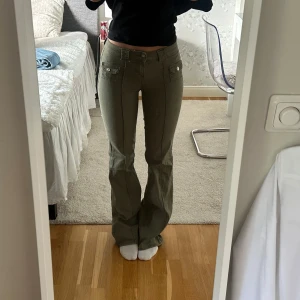 Olivgröna bootcut byxor med fickor - Snygga olivgröna byxor med bootcut och dekorativa fickor framtill med knappar. Byxorna har låg midja och markerade sömmar längs benen för en cool look. Perfekta för dig som gillar en avslappnad men trendig stil.