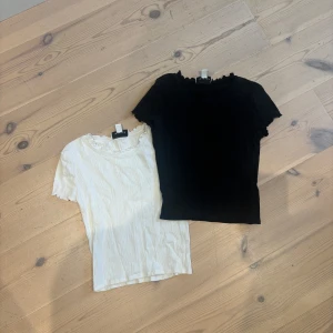 2 ribbade toppar med volangkant - Två söta ribbade toppar  i svart och vit. Båda har kort ärm, volangkant vid halsringning och ärmslut samt en lätt figurnära passform. Perfekta att styla med jeans eller kjol för en enkel men trendig look.