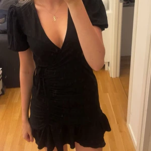 Svart omlottklänning med volang - Säljer min jättefina klänning då den aldrig kommer till användning! Nyskick, inga defekter. Super skön och luftig, skriv vid fler frågor💗