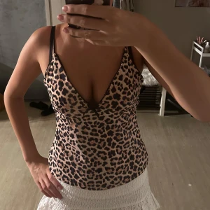 Leopardmönstrad linnetopp med rosett - Säljer en leopardmönstrad topp med smala svarta axelband och djup v-ringning. Toppen har en söt svart rosett framtill och dekorativ spetskant längs urringningen. Materialet är mjukt och följsamt, perfekt för dig som vill sticka ut med ett djuriskt mönster.