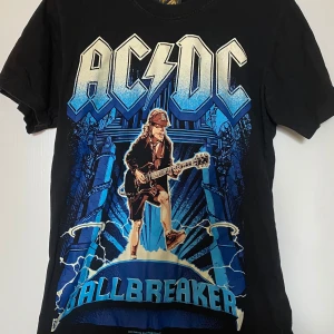 Svart AC/DC Ballbreaker t-shirt S - Svart t-shirt med stor AC/DC Ballbreaker-tryck i blått och vitt framtill. Klassisk rund hals och korta ärmar. Gjord i 100% bomull, perfekt för dig som gillar rock och vill sticka ut med en fet bandtröja. Frakt tillkommer