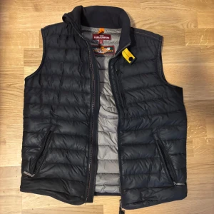 Svart dunväst från Parajumpers - Säljer en svart quiltad dunväst från Parajumpers med grå insida och gul detalj vid kragen. Västen har två dragkedjefickor och hög krage. Superlätt och perfekt för lager-på-lager. Klassisk och stilren design med snygga detaljer.