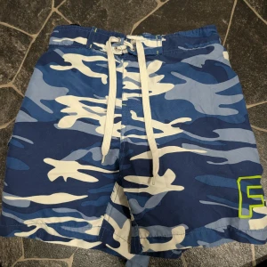 Blå camo badshorts från Funk stl 140 - Snygga blå camo badshorts från Funk med vita och ljusblå detaljer. Shortsen har snörning i midjan och ett tryckt F i neonfärg på benet. Perfekta för stranden eller poolen, tillverkade i 100% polyester för snabb torkning.
