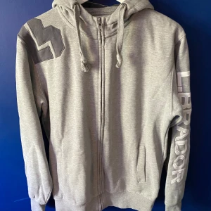 Grå hoodie från L.Brador med tryck - Snygg grå hoodie från L.Brador med dragkedja, huva med snören och stort broderat logotyp-tryck längs ena ärmen. Mjuk bomullsblandning och ribbade muddar. Perfekt för chill dagar eller när du vill ha en clean streetstyle.