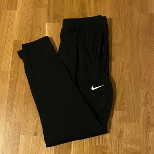 Svarta Nike Dri-FIT träningsbyxor  - Svarta träningsbyxor från Nike med Dri-FIT-teknologi. Byxorna har elastisk midja, dragkedja vid fickorna och en vit Nike-logga på benet. Perfekta för sport eller chill, tillverkade i lätt och snabbtorkande syntetmaterial.