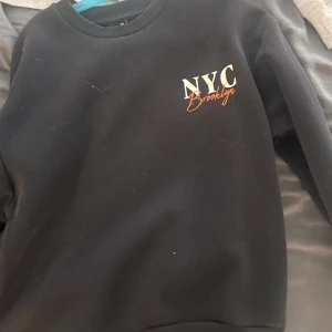 Svart sweatshirt med Brooklyn-tryck - Svart sweatshirt med tryck på både fram- och baksidan. På bröstet står det 'NYC Brooklyn' i vitt och orange, och på ryggen finns ett stort tryck med texten 'Brooklyn New York City USA' och flera gatunamn. Klassisk rund hals och långa ärmar.