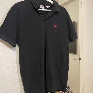 Svart pikétröja från Levi's - Klassisk svart pikétröja från Levi's med korta ärmar och liten röd logotyp på bröstet. Tröjan har krage och knäppning framtill. Perfekt för en clean och stilren look.