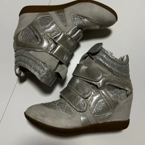 isabel marant - Unika sneakers i silver med kilklack och coola detaljer. Skorna har flera breda kardborreband, glittriga meshpartier och inslag av mocka. Perfekta för dig som vill sticka ut med en futuristisk vibe och snygg höjd. Snyggt kontrasterande sula i brunt. Sor är inspirerad av isabel marant. Pris går att diskutera❤️