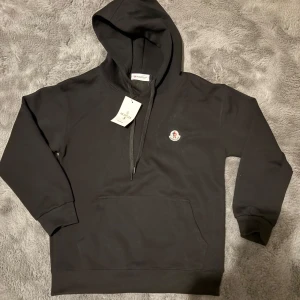 Svart hoodie från Moncler - Säljer en svart hoodie från Moncler med klassisk logotyp på bröstet. Tröjan har huva med dragsko och en stor magficka framtill. Perfekt för en avslappnad och stilren look.
