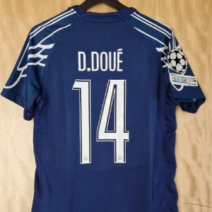PSG Champions League tröja - Blå PSG fotbollströja med vita detaljer, tryck D. Doué #14 på ryggen och Champions League-märke på ärmen. Jordan-logga och Paris Saint-Germain-emblem på bröstet, samt Qatar Airways som sponsor. Tillverkad i lätt och ventilerande material.