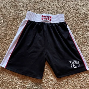 Svarta boxningsshorts från Paffen Sport XS - Boxningsshorts från Paffen Sport i svart med vita sidor och röda detaljer. Bred, elastisk midja med logga framtill och PS-brodyr på benet. Lätta och luftiga, perfekta för träning eller match.                     Nypris 299kr! 