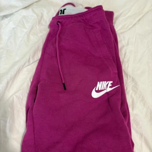 Rosa mjukisbyxor från Nike - Snygga rosa mjukisbyxor från Nike med vit logga på benet och praktisk snörning i midjan. Perfekta för chill eller träning, med mjukt material och bekväm passform.