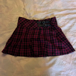 Rutig kortkjol med spännen - Supersnygg rutig kortkjol i mörkrött och svart med två coola spännen i midjan. Kjolen har en klassisk kiltstil och är perfekt för dig som vill sticka ut med en edgy vibe. Passar grymt till boots och en oversized tröja.