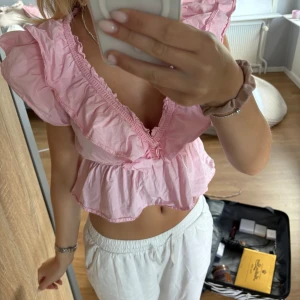 Rosa volangblus med v-ringning - Supersöt rosa blus med djup v-ringning och volangdetaljer längs axlar och hals. Modellen är croppad och har en luftig passform, perfekt för varma dagar. Tillverkad i lätt bomullstyg som känns skönt mot huden.