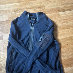 Marinblå kofta från Replay med dragkedja - Snygg marinblå kofta från Replay med hel dragkedja framtill och ribbad krage. Ärmarna är långa och ena ärmen har en cool ficka med dragkedja. Materialet är stickad bomull och passformen är normal. Perfekt för dig som gillar stilrena detaljer och funktion.