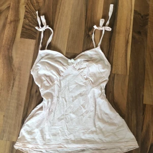 Ljusrosa linnetopp med rosett - Supersöt ljusrosa linnetopp med smala axelband som knyts på axlarna och en liten rosett framtill. Toppen har en feminin v-ringning och är tillverkad i mjuk bomull, perfekt för varma dagar.