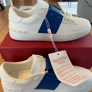Valentino Garavani Open sneakers med blå detalj - Säljer ett par vita Valentino Garavani Open sneakers med bred blå rem över sidan. Skorna har rund tå, platt sula och ikoniska nitar baktill. Märkesdetaljer på plösen och sula. Levereras med originalkartong och tillbehör.