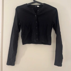 Svart croppad tröja med knappar - Säljer en svart croppad tröja med långa ärmar och rund halsringning. Tröjan har knäppning framtill med svarta knappar och är tillverkad i ett mjukt bomullsmaterial. Perfekt att styla över ett linne eller t-shirt.