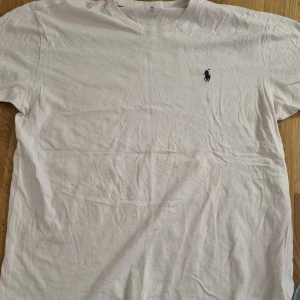 Vit t-shirt från Ralph Lauren - Klassisk vit t-shirt från Ralph Lauren med liten broderad logga i mörkblått på bröstet. T-shirten har rund hals och korta ärmar, perfekt för en clean och stilren look.