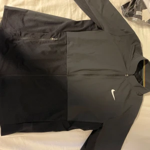 Svart träningsjacka från Nike - Säljer en svart träningsjacka från Nike med dragkedja framtill och klassisk vit logga på bröstet. Jackan har lång ärm och är tillverkad i ett lätt syntetmaterial som andas. Perfekt för träning eller sportiga dagar.