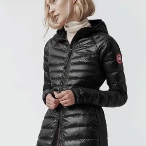 Canada Goose Hybridge Jacket - Säljer en svart, lång och figurnära dunjacka från Canada Goose med huva och dragkedja. Jackan har diskret logga på ärmen och justerbara knappar i midjan. Perfekt för kyliga dagar. Passar perfekt till våren/hösten. Köpt i NK så den är äkta såklart  och mycket sparsamt använd.Jag säljer den för att jag inte använder den då det inte är min stil längre. Passar xs/s .Nypris ligger runt på 11000kr 