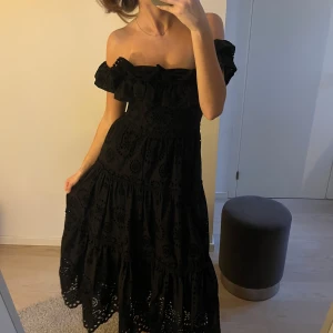 Svart offshoulder långklänning med volang - Säljer en svart långklänning med offshoulder-modell och volang upptill. Klänningen har broderade detaljer och vacker hålbrodyr i tyget, vilket ger en romantisk känsla. Perfekt för sommarens tillfällen.