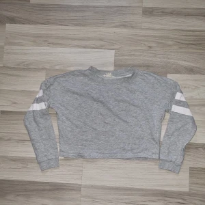Grå croppad sweatshirt från H&M - Grå croppad sweatshirt från H&M med långa ärmar och vita ränder på båda ärmarna. Tröjan har en enkel och sportig look, perfekt för en avslappnad stil. Mjuk bomullskänsla och rund halsringning.