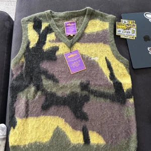 Camo mönstrad mohairväst från Corteiz - Säljer en camo mönstrad stickad väst från Corteiz i gröna, bruna och gula toner. Västen är v-ringad och har en mjuk, luddig känsla tack vare mohairblandningen. Perfekt för dig som vill sticka ut med en unik look.