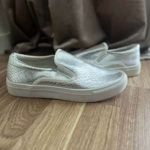 Silvriga slip-on sneakers - Säljer ett par silvriga slip-on sneakers från Skopunkten med reptilmönstrad yta och vit platt sula. Skorna är i syntetmaterial och har en rund tå. Perfekta för dig som vill sticka ut med en glittrig touch till vardagslooken.