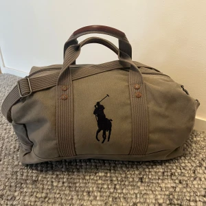 Olivgrön weekendbag från Polo Ralph Lauren - Snygg olivgrön weekendbag från Polo Ralph Lauren med klassisk logga framtill. Väskan har både handtag i brunt skinn och justerbar axelrem. Perfekt storlek för en kort resa eller gymmet. Stängs med dragkedja och har robust canvasmaterial. ‼️Nypris ca 3200kr. Mitt pris: 1450kr men pris kan även sänkas‼️