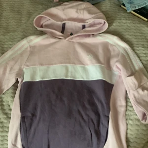 Ljusrosa och lila hoodie från Adidas - Säljer en snygg hoodie från Adidas i ljusrosa och lila med vit rand över bröstet. Tröjan har huva, långa ärmar och klassiska Adidas-ränder på axlarna. Perfekt för en sportig och avslappnad stil. Meddela om du vill veta hur den är på!💗