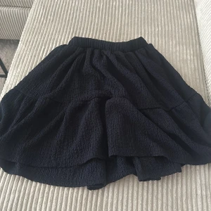 Svart volangkjol med resårmidja - Säljer en svart kjol med volanger och resår i midjan. Kjolen har en kort längd och är tillverkad i ett lätt, strukturerat tyg som ger ett fint fall. Perfekt att styla med en enkel topp eller stickad tröja.