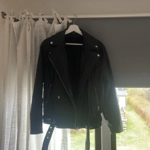 Svart skinnjacka med dragkedjor och bälte - Säljer en svart skinnjacka med silvriga dragkedjor, knappar och bälte i midjan. Jackan har långa ärmar och bred krage. Supersnygga att också ta ur bältet och knyta de i en knut runt midjan. Inga defekter.💕💕