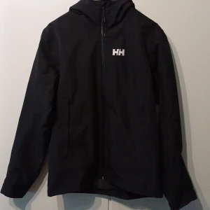 Svart vindjacka från Helly Hansen - Svart skaljackafrån Helly Hansen med dragkedja framtill och logga på bröstet. Jackan har huva och är tillverkad i slitstarkt syntetmaterial med Helly Tech-skydd. Perfekt för blåsiga dagar och utomhusaktiviteter.