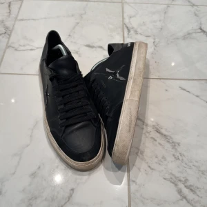 Svarta sneakers från Axel Arigato med broderad fågel - Svarta sneakers i skinn från Axel Arigato med broderad fågel på sidan och guldfärgad logga. Skorna har svarta snören, rund tå och vit platt sula. Perfekta för en stilren och modern look. Box samt dust bag medföljer. Nypris 2660kr mitt pris 899kr💯