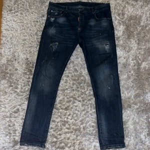 Mörkblå jeansbyxor från Dsquared2 - Säljer ett par mörkblå jeansbyxor från Dsquared2 med slitningar och färgstänk för en cool, använd look. Byxorna har klassisk femficksdesign, knappgylf och röd Dsquared2-tag vid gylfen. Perfekta för dig som gillar streetstyle. Pris går att diskutera ✅