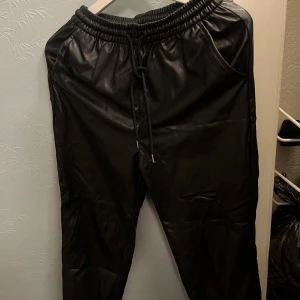 Svarta syntetbyxor från H&M Divided - Säljer ett par svarta byxor från H&M Divided med resår och dragsko i midjan. Byxorna har en glansig finish som liknar skinn och är tillverkade i syntetmaterial. Perfekta för en trendig och avslappnad look.