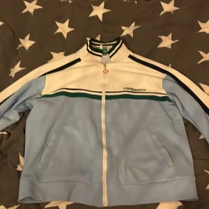 Casablanca tennisjacka i blått och vitt  - Säljer en snygg tennisjacka från Casablanca i ljusblått och vitt med gröna och svarta detaljer. Jackan har dragkedja framtill, ståkrage med randigt mönster och två fickor. Perfekt för en sportig look.