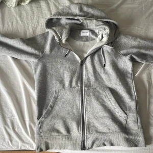 Grå hoodie med dragkedja från Jack & Jones - Säljer en klassisk grå hoodie från Jack & Jones i storlek S, använd cirka 1-3 gånger enbart. Tröjan har dragkedja framtill, två stora fickor och justerbar huva med snören. Perfekt för en avslappnad stil och enkel att matcha med det mesta.