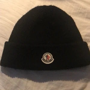 Moncler mössa  - Säljer en svart stickad mössa från Moncler med uppvikt kant och klassisk Moncler-logga framtill. Perfekt för kyliga dagar och enkel att matcha med olika outfits.