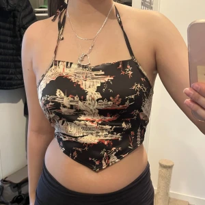 Svart halterneck topp med asiatiskt mönster - Säljer en svart topp i siden med mönster i beige och rött från Urban outfitters. Toppen är croppad och knyts både i nacken och ryggen. Använd en gång.