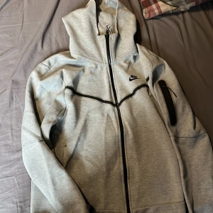 Ljusgrå hoodiejacka från Nike - Säljer en ljusgrå hoodiejacka från Nike med hel dragkedja och svarta detaljer. Jackan har huva, lång ärm och en ficka med dragkedja på ena ärmen. Perfekt för träning eller chill.