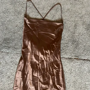 Brun satin klänning med smala axelband från H&M - Säljer en brun satin klänning från H&M med smala axelband och figurnära passform. Klänningen har en elegant glansig yta och är perfekt för dig som gillar en stilren look. Lätt och följsam modell som ger en snygg siluett. Mycket bra skick då det aldrig kommit till användning!