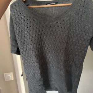 Grå stickad T-shirt från Filippa K - Mysig grå stickad t-shirt från Filippa K med korta ärmar och rund halsringning. Tröjan har ett diskret hålmönster upptill och bredare ribbstickning nedtill och vid ärmslut. Perfekt nu till våren och sommaren. Hör av er vid frågor 🥰