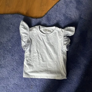 Vit topp med volangärmar från Kappahl - Säljer en söt vit topp från Kappahl med volangdetaljer på axlarna. T-shirten har rund halsringning och är tillverkad i mjuk bomull. Perfekt för en fräsch och stilren look.