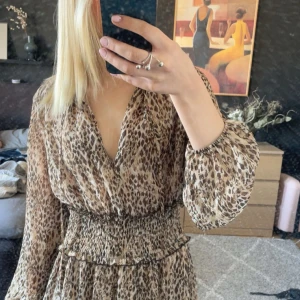 Leopardmönstrad klänning från Zara - Säljer en snygg leopardmönstrad klänning från Zara med v-ringning och långa ärmar. Klänningen har smock i midjan som ger en fin passform och ett luftigt fall. Perfekt för dig som gillar djurmönster och vill sticka ut.