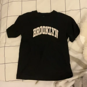 Svart t-shirt med BROOKLYN-tryck från H&M - Svart t-shirt från H&M med vit text 'BROOKLYN' tryckt på bröstet. Klassisk rund hals och korta ärmar. Perfekt för en avslappnad streetstil och enkel att matcha med jeans eller shorts. Är som ny, använd 2-3 gånger men har inte prislapp kvar.